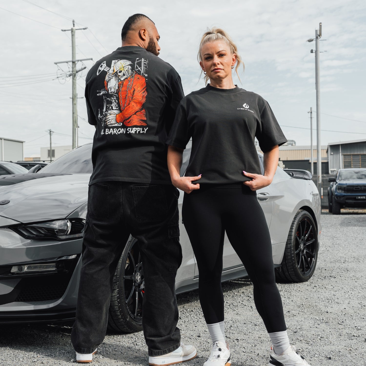 Two people wearing black t-shirts standing next to a gray car in an outdoor setting.