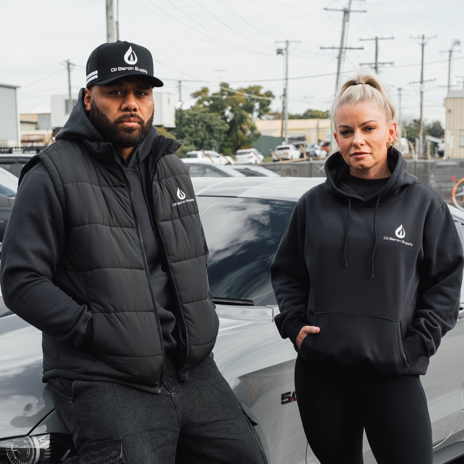 Two people wearing black hoodies with an oil baron supply logo standing next to a car.