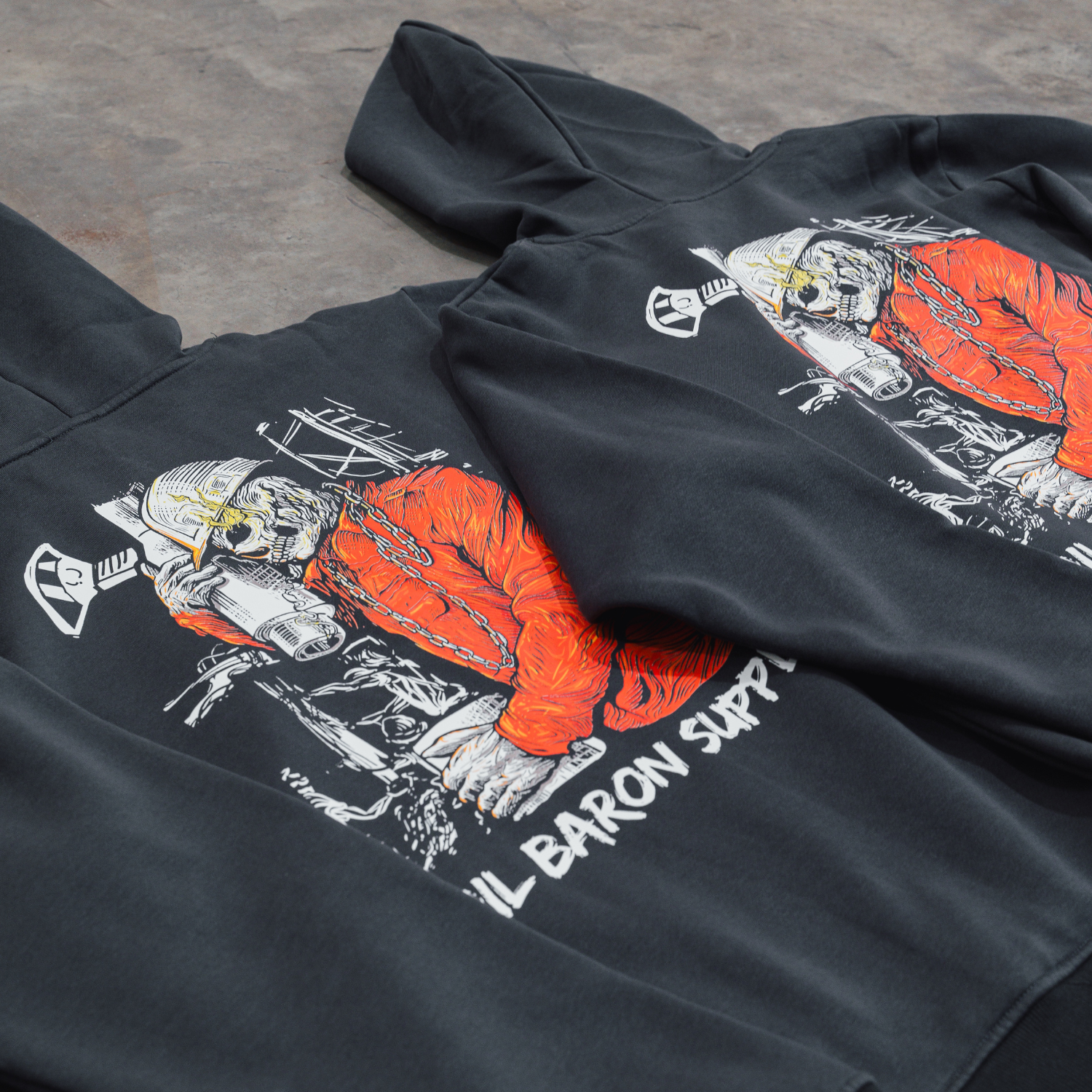 Black hoodie with a Roughneck design of a skeleton in an orange outfit on a concrete floor.