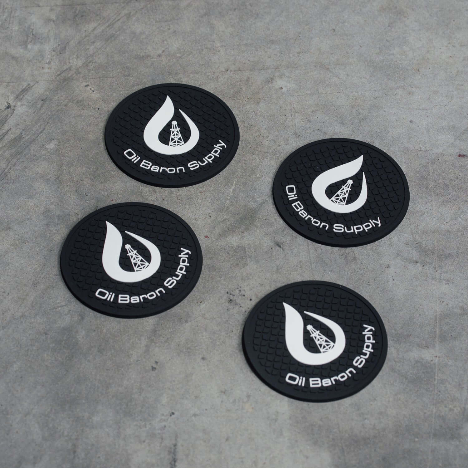 Four black coasters with 'Oil Baron Supply' logo on a concrete surface