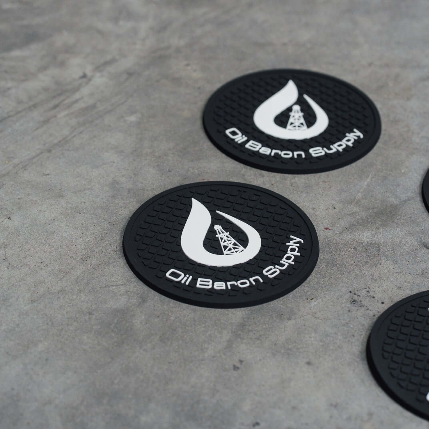Black coasters with 'Oil Baron Supply' logo on a concrete surface