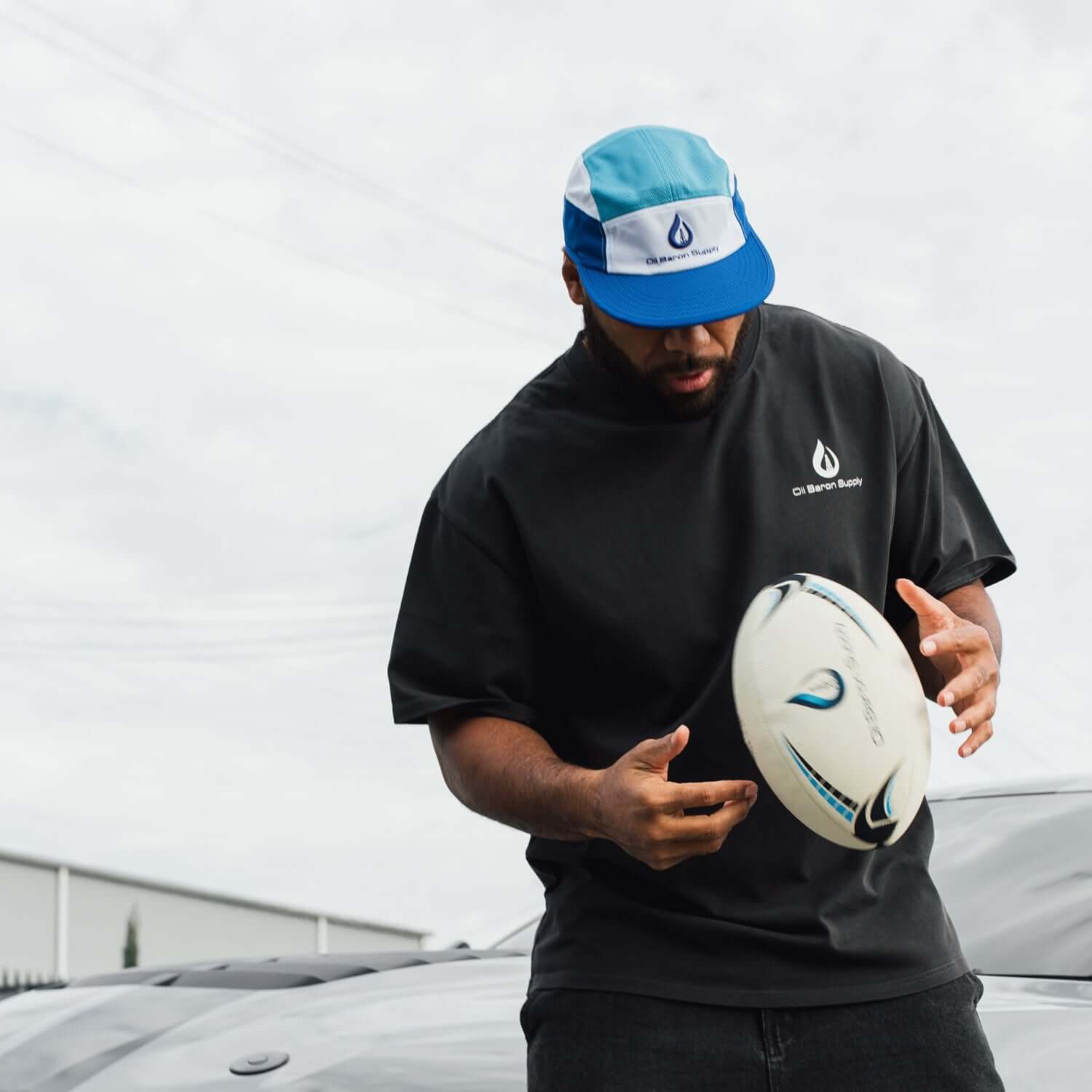 Person holding a rugby ball wearing a black t-shirt and blue and white cap with a logo.